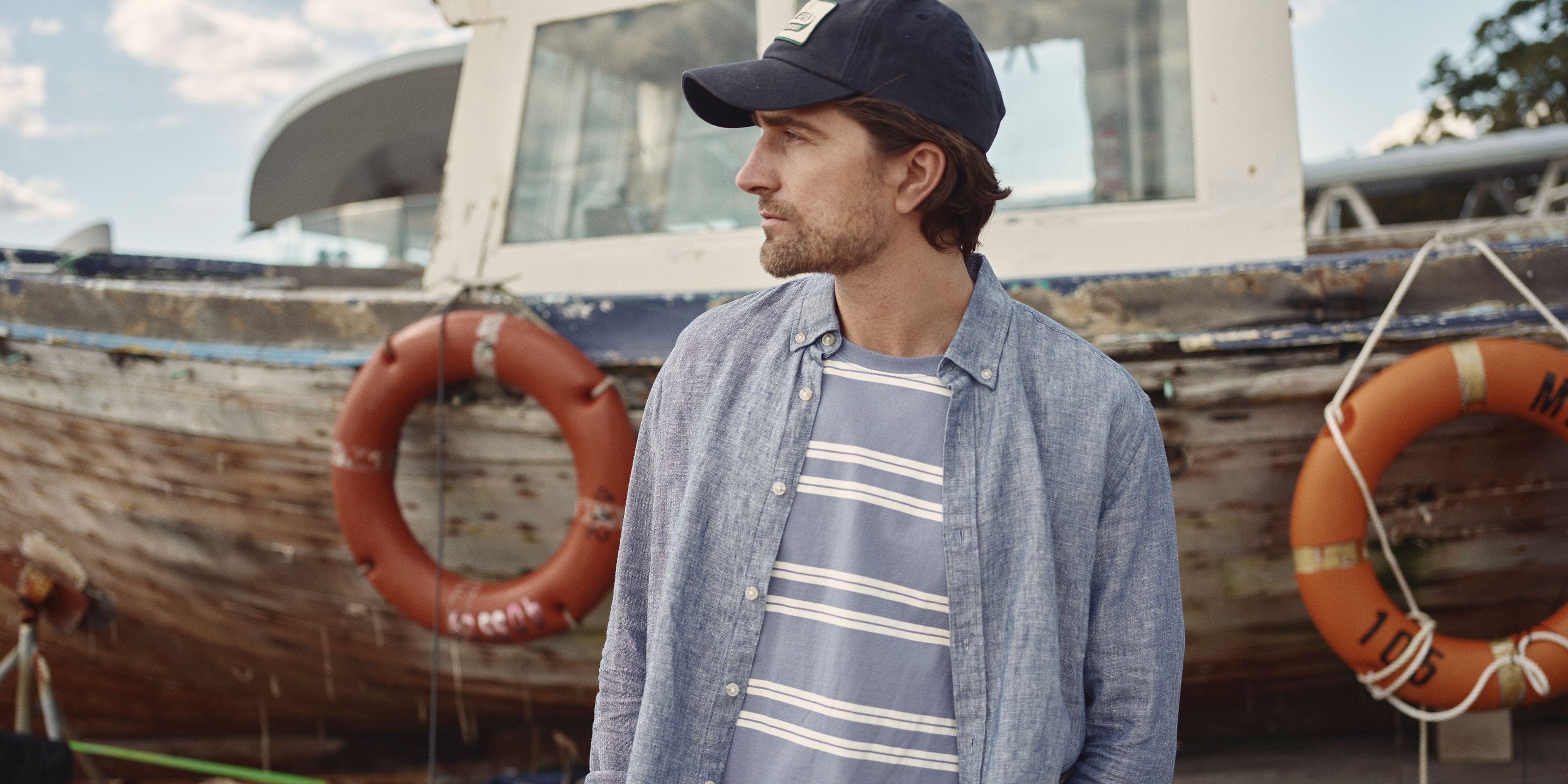 A man in a navy cap, blue striped shirt, and light blue button-up stands in front of an old wooden boat with orange life rings. He looks to the right, and the scene appears outdoors near the water.