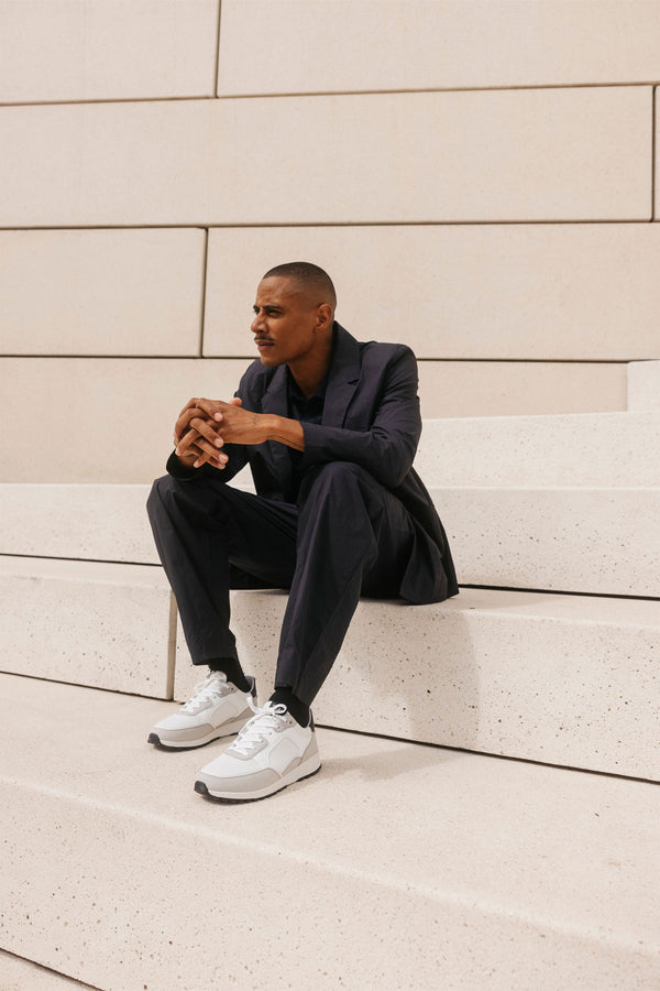 A man in a dark suit and Clae Joshua vegan trainers sits on wide, white concrete steps outdoors, gazing to the side with hands clasped, framed by large smooth stone blocks.