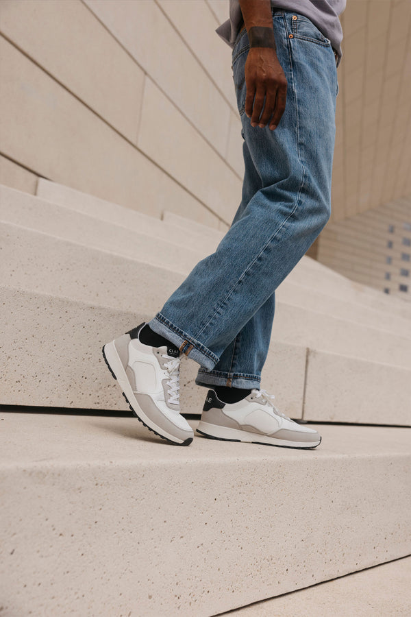 A person in blue jeans and Clae - Joshua sustainable white sneakers stands on light concrete steps, left hand at their side, with a modern beige building in the background.