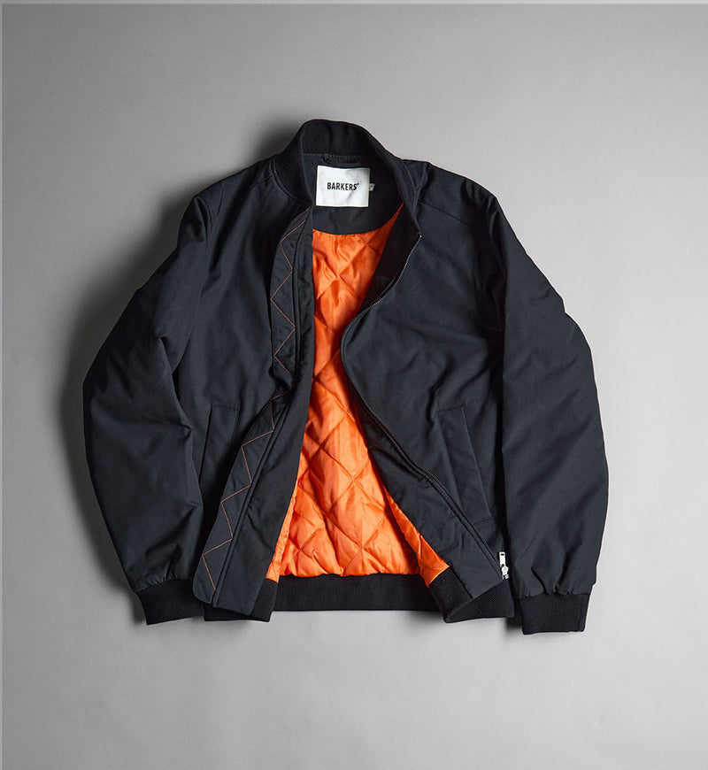 Black NZ bomber jacket with orange quilted lining, laid flat on a gray surface.