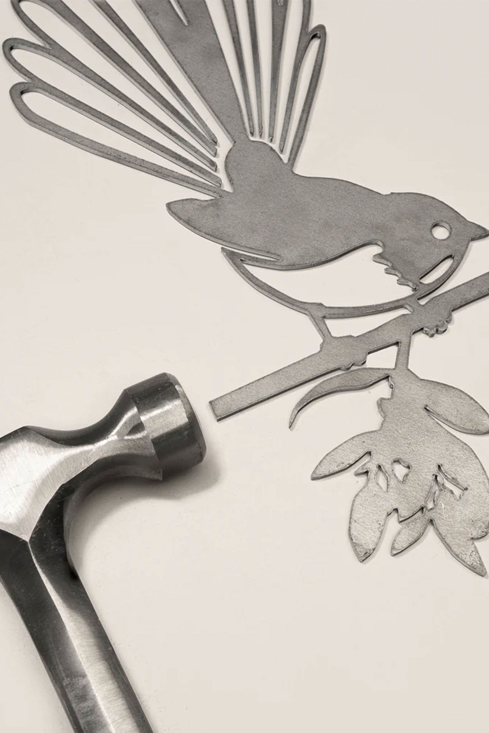 The Other brand "Piwakawaka / Fantail" Corten steel bird cutout, with a silhouette of the bird on a floral branch, rests on a flat surface beside a silver hammer.