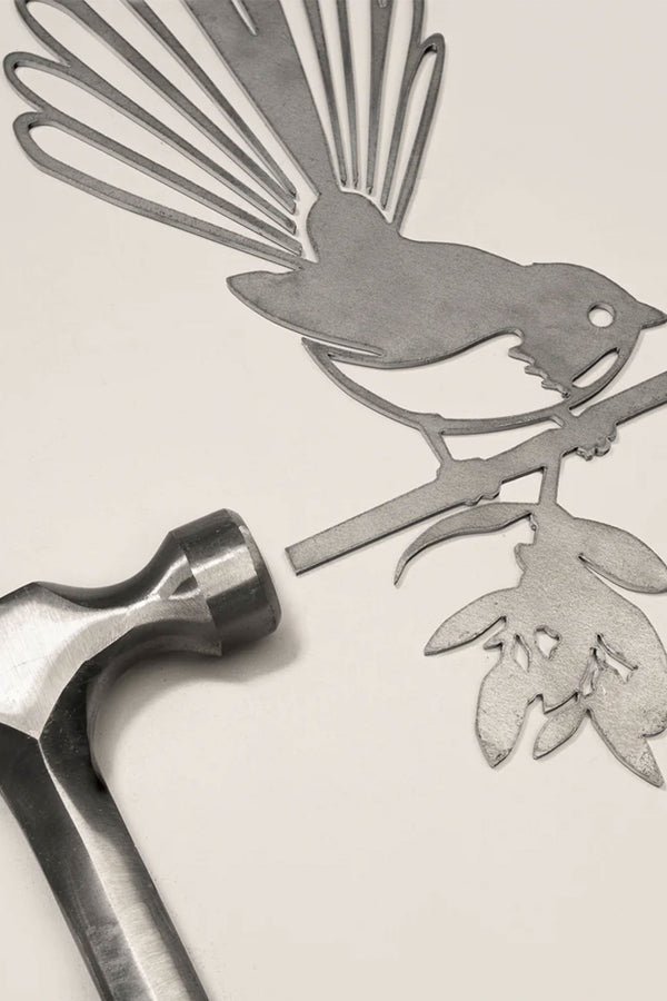 The Other brand "Piwakawaka / Fantail" Corten steel bird cutout, with a silhouette of the bird on a floral branch, rests on a flat surface beside a silver hammer.