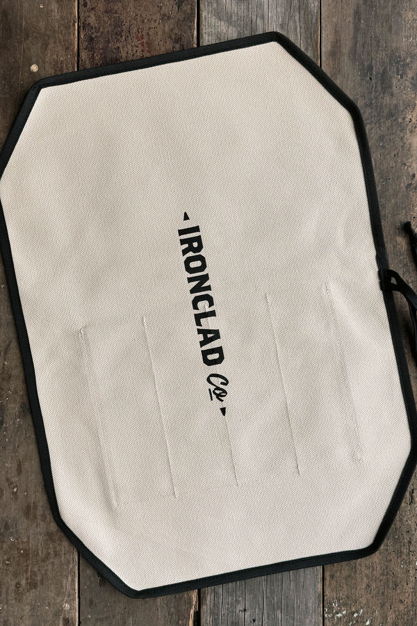 A white, octagonal IronClad apron with black trim and logo lies flat on a rustic wooden surface beside an IronClad Ghost Knife Roll.