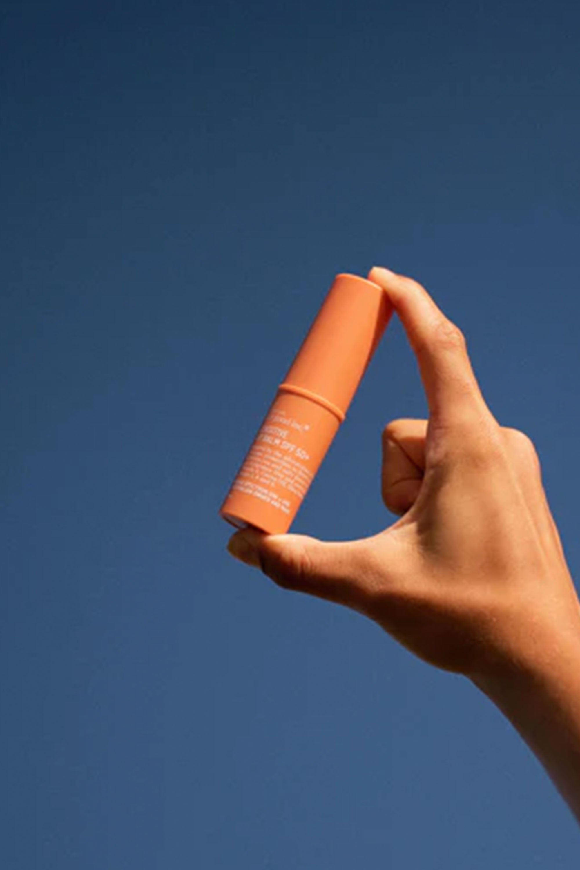 A hand holds Other Sensitive Lip Balm SPF50+ 10ml against a clear blue sky.