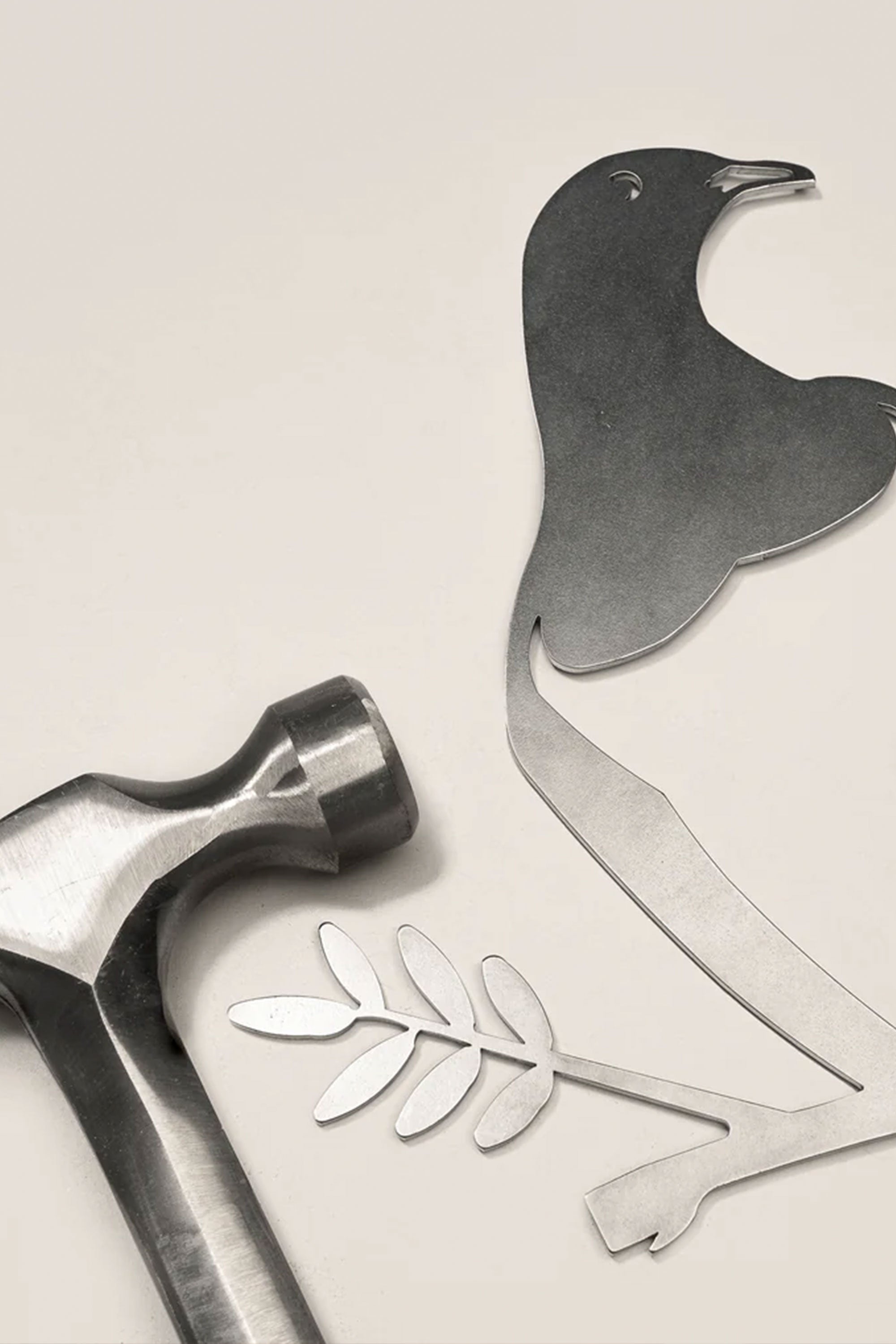 A metal hammer lies next to cut-out pieces forming the Other brand Metal Birds Kereru / Woodpigeon, a Corten steel bird with an olive branch, inspired by native New Zealand art, all displayed on a light surface.