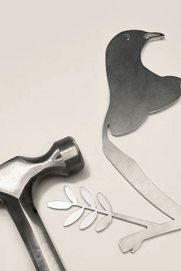 A metal hammer lies next to cut-out pieces forming the Other brand Metal Birds Kereru / Woodpigeon, a Corten steel bird with an olive branch, inspired by native New Zealand art, all displayed on a light surface.